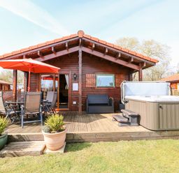 A wooden lodge with a deck featuring a hot tub table with chairs and an orange umbrella at Bittern Lodge in Tattershall Lakes Country Park