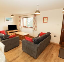 A living room with two sofas, red cushions, a wooden coffee table, television on a stand, windows with floral curtains, and a wooden door at Bodneithior in Llanerchymedd