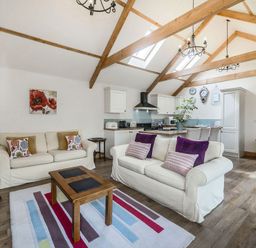 An open plan living room and kitchen with wooden beams on the ceiling and sofas with cushions at Warbrook in Bradworthy