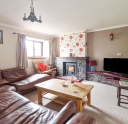 A living room with a brown leather sectional sofa a wooden coffee table a rocking chair a stone fireplace with floral wallpaper a TV on a stone shelf and a wooden chair at Haulfryn in Bethesda