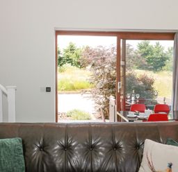 A living room with a dark brown leather sofa and cushions near a large glass sliding door with red chairs at Casterbridge Upwey near Weymouth