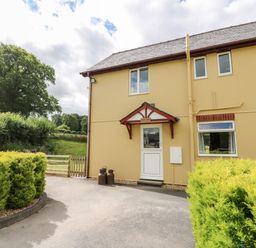 The exterior of a yellow two-story house with a white door and red canopy surrounded by green bushes at Red Kite Cottage in Howey near Llandrindod Wells