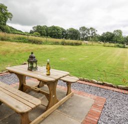 A wooden picnic table with a lantern wine bottle and two glasses on a patio overlooking a fenced grassy field with sheep at Red Kite Cottage in Howey near Llandrindod Wells