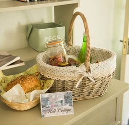 A wicker basket with a jar of granola and a green bottle next to a basket with bread on a dresser at Mill Cottage Hanley William near Tenbury Wells