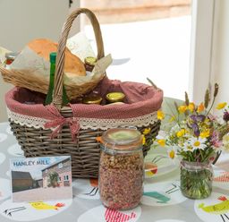 A basket with bread jars and a bottle on a table with a jar of granola flowers and a postcard at Stable Cottage in Hanley William near Tenbury Wells