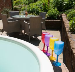 An outdoor patio with a round table and four wicker chairs a bottle and glasses on the table and colorful plastic wine glasses on the edge of a round hot tub at Stable Cottage Hanley William near Tenbury Wells