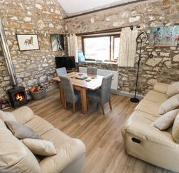 A living room with beige sofas a wooden dining table with grey chairs a wood burning stove and stone walls at Corner Cottage in Broad Haven