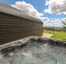 An outdoor hot tub next to a wooden building with a view of a grassy field and hills at Valley View in Rhayader