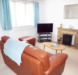 A living room with a brown leather sofa, light blue curtains, a TV on a stand, a wooden coffee table with magazines, and a stone fireplace with a small stove at Kates Cottage in Bourton-on-the-Water