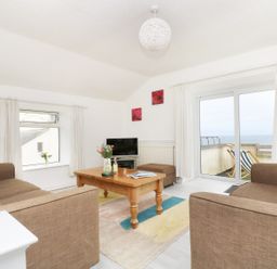 A living room with brown sofas a wooden coffee table a rug and a balcony with deck chairs at The Coach House in Sennen near St Just