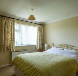 A bedroom with a bed with a metal headboard bedside tables with lamps a window with curtains and a chair in Skylark Weybourne