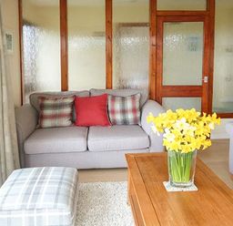 A living room with a sofa with cushions a wooden coffee table with a vase of yellow flowers a plaid ottoman and a wooden cabinet with a lamp at Pine Lodge in Culbokie