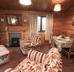 A living room with floral and striped armchairs a dining table set with dishes curtains on the window and wood paneling at Sycamore Lodge in Liverton near Danby
