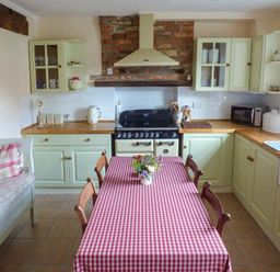 A kitchen with a wooden table covered with a red and white checkered tablecloth surrounded by wooden chairs a couch with cushions green cabinets a stove and a sink at Woodside Cottage in Brompton-by-Sawdon