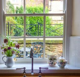 A kitchen window with a sink and faucet with a white pitcher holding flowers and ceramic containers on the windowsill with a view of an outdoor patio with metal chairs and greenery at Woodside Cottage in Brompton-by-Sawdon