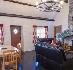 A living room with a wooden dining table and chairs a stone fireplace with firewood and a wood stove black leather sofas and floral curtains at The Well House in Kinvara County Galway