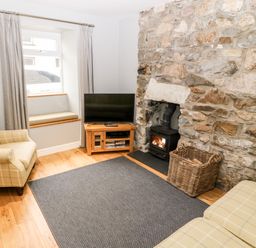 A sitting room with a TV on a wooden stand a stone fireplace with a wood burner and two checked armchairs at Haddef in Nefyn