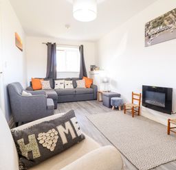 A living room with a gray sofa and armchair two wooden chairs a wall-mounted fireplace and a window with curtains at Sealyons Fairbourne near Dolgellau