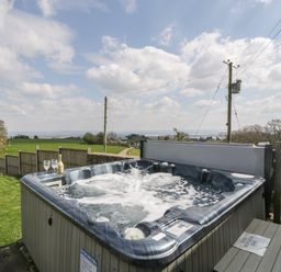 An outdoor hot tub with two glasses and a bottle on the edge overlooking a grassy field and a rural landscape at Moors Farm in Littledean
