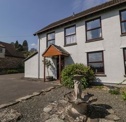 A white two-story house with brown framed windows and a small porch next to a stone fountain and bushes at Moors Farm in Littledean