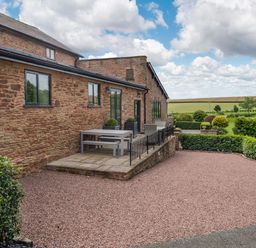 An outdoor patio area with stone tiles and a table with benches next to a brick building with a gravel driveway and manicured hedges at Teal Barn Annexe in St Weonards