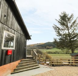 An exterior view of a building with wooden siding steps and a railing overlooking a rural landscape with a tree and fenced area at Middle Hivron in Bleddfa