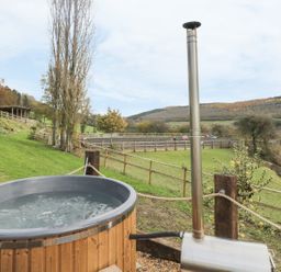 An outdoor hot tub with a wood stove in a fenced grassy area with hills and trees at Middle Hivron in Bleddfa