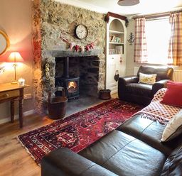 A living room with a stone fireplace and wood stove two leather sofas a wooden table with a lamp and flowers and a red patterned rug at Bwthyn Megan in Llangaffo near Newborough