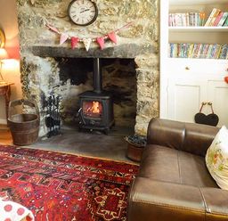 A living room with a stone fireplace wood burning stove a brown leather sofa and a bookcase filled with books and DVDs at Bwthyn Megan in Llangaffo near Newborough