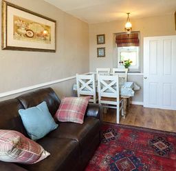 A seating area with a brown sofa and dining table with white chairs near a window at Bwthyn Megan in Llangaffo near Newborough