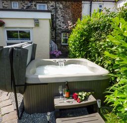 An outdoor hot tub with water flowing into it surrounded by green bushes and a small table with wine glasses and flowers at Bwthyn Megan in Llangaffo near Newborough
