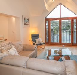A living room with a leather sofa, glass coffee table, armchair, floor lamp, and large windows overlooking a hillside at The Creekside in Looe