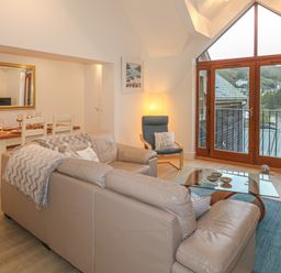 A living room with a beige leather sectional sofa a glass coffee table a dining table with chairs and large windows showing a view outside at The Creekside in Looe
