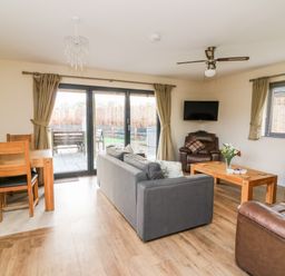 A living room with a gray sofa, wooden coffee table with flowers, brown leather chairs, dining table with chairs, ceiling fan, and sliding glass doors at Caban Nant Ddu in Rhayader