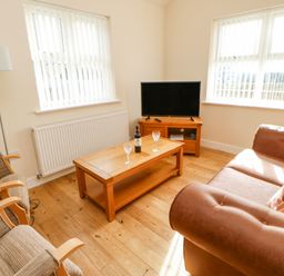 A living room with a brown sofa two armchairs a wooden coffee table with a bottle and two glasses a wooden TV stand and a television at Wansbeck Longframlington near Alnwick