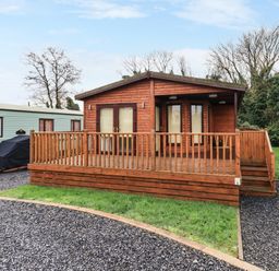 A wooden cabin with a front porch and railing surrounded by gravel and grass at Roe Deer Lodge in Castlewigg near Whithorn