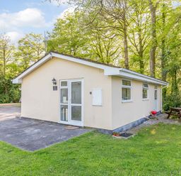 A small cream-colored building with glass door and windows surrounded by grass and trees at Brook Cottage in Rosecraddoc Lodge near Liskeard