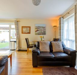 A living room with leather sofas a wooden dining table and chairs next to a glass door and large windows with curtains at Brook Cottage Rosecraddoc Lodge near Liskeard