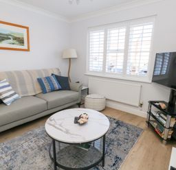 A living room with a grey sofa with cushions a round coffee table a floor lamp a TV on a stand and a window with white shutters at Captains Retreat in Christchurch