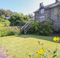 A garden with a lawn and yellow flowers next to a stone house with wooden stairs at Snowdon View in Harlech