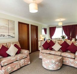 A living room with floral sofas and matching ottoman with red and cream cushions at Chy Lowenna Rosecraddoc Holiday Village near Liskeard