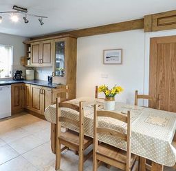 A kitchen with wooden cabinets dining table with tablecloth and chairs and flowers in a vase at Simdda Wen Cottage in Llanddona near Beaumaris