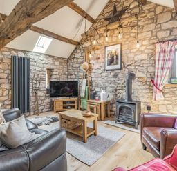 A living room with stone walls wooden beams a wood-burning stove a leather sofa and armchairs a television and wooden furniture at Berts Cottage in Heights near Appleby-in-Westmorland