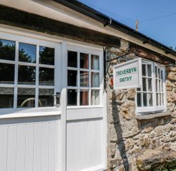 Exterior of a stone building with white framed windows and doors with a sign reading Treverbyn Smithy in St Neot