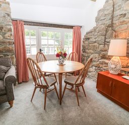 A dining area with a round wooden table and four wooden chairs near a window with red curtains at Treverbyn Smithy in St Neot