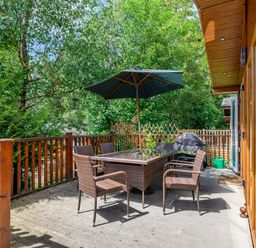 An outdoor wooden deck with a glass dining table, six wicker chairs, a large umbrella, a barbecue grill, and surrounding trees at Claife View Lodge in Troutbeck Bridge