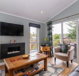 A living room with a wall mounted TV fireplace wooden table armchair and sliding glass doors at Claife View Lodge in Troutbeck Bridge