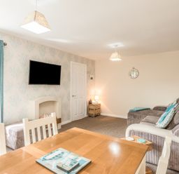 A living room with a dining table, chairs, a wall-mounted TV, sofa, small side table with lamp, and patterned wallpaper at Rock Rose in Beadnell