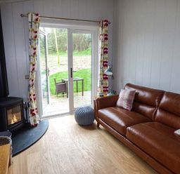 A living room with a brown leather sofa two pillows a wood burning stove a sliding glass door with curtains and a small outdoor table and chair at Shepherd Lodge in Allithwaite near Grange-over-Sands