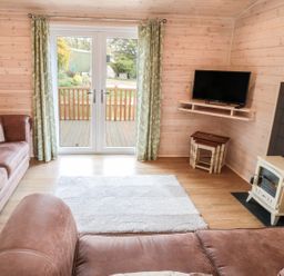 A living room with brown sofas a white rug a wall-mounted TV a small stove and glass doors with green curtains at Thorntree Lodge in Rowsley